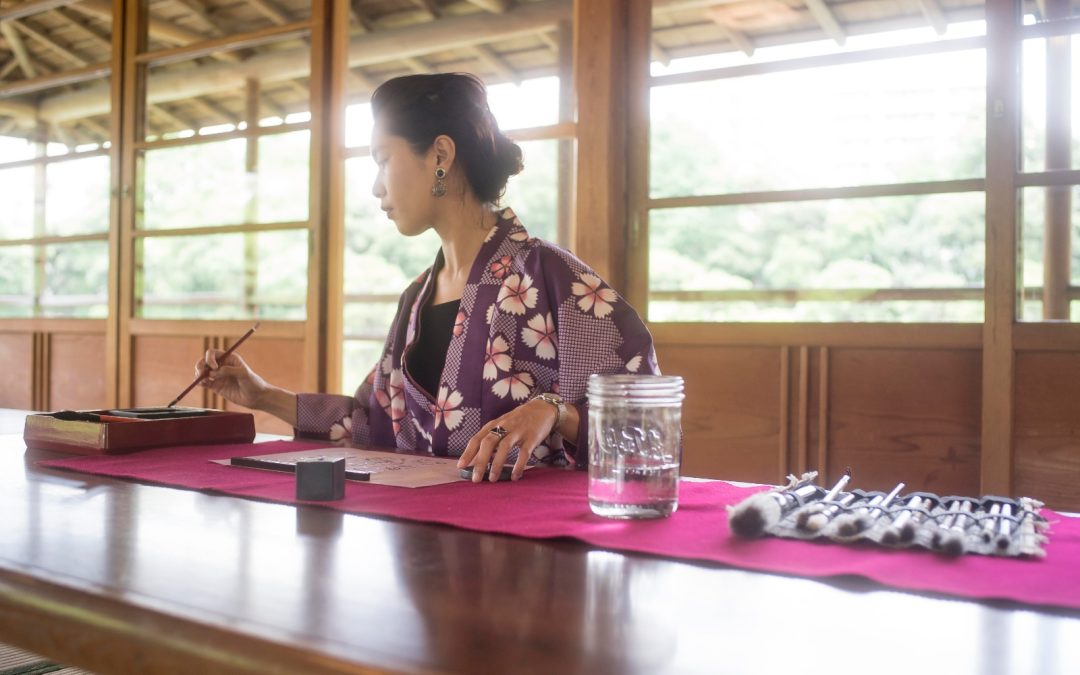 Quiet Vitality China: woman in traditional robe practicing calligraphy at a wooden table with brushes and ink in a calm room.